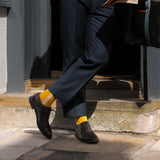 Man leaning against a wall wearing a navy suit, brown shoes and mustard Andover men's luxury socks by Peper Harow