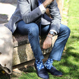 Man sitting on edge of fountain wearing jeans, a check blazer, navy shoes and navy Triangle men's luxury socks by Peper Harow